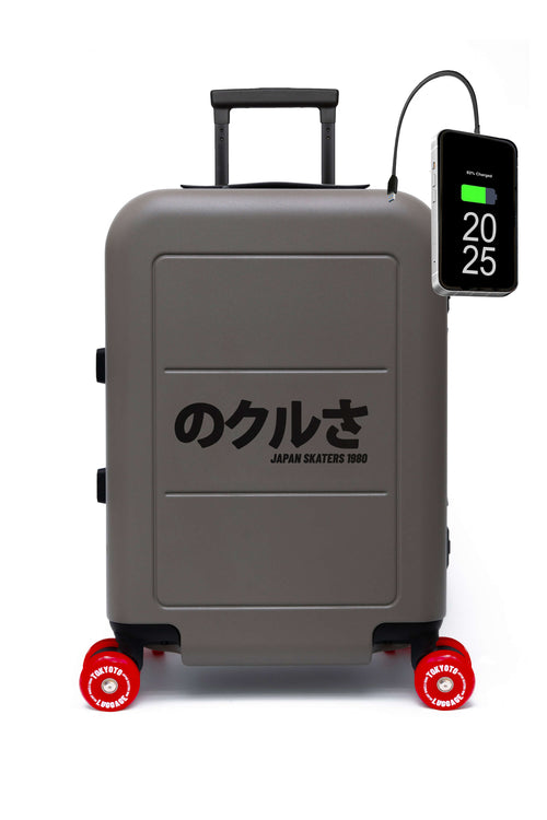 A matte gray hard-shell skate wheels suitcase with a black telescoping handle and four wheels, featuring the black Japanese text JAPAN SKATERS on the front. A black smartphone power bank is connected to its side