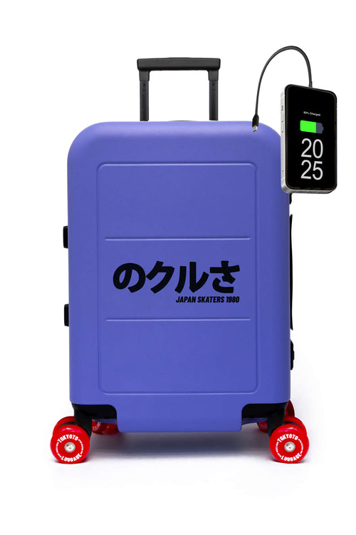 Ein lavendelblauer Hartschalenkoffer mit Skateboardrollen, roten Rädern und schwarzem japanischem Text auf der Vorderseite: „のクルさ JAPAN SKATEERS 2020“. Ein Smartphone-Akkuladegerät ist seitlich am Koffer eingesteckt, vor einem schlichten weißen Hintergrund.